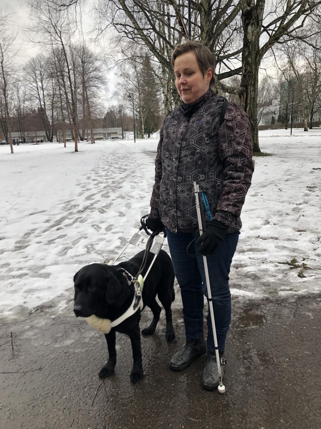 Susanna ja labradorinnoutaja opaskoira Ape seisovat vähä lumisessa puistossa. Apella on päällä valkoiset valjaat, joista Susanna pitää kiinni oikealla kädellään. Susannalla on päällään siniset farkut ja mustavalkoinen talvitakki. Vasemmassa kädessä Susannalla on valkoinen keppi.